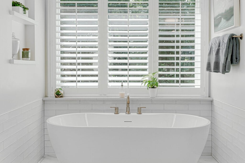 A bright, modern bathroom featuring a freestanding oval bath, sitting beneath three windows covered with classic white plantation shutters.