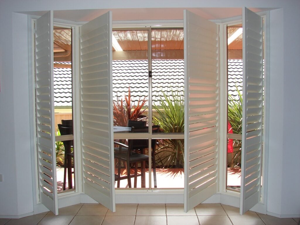 A window covered by six white plantation shutters, with the two central panels fully opened.