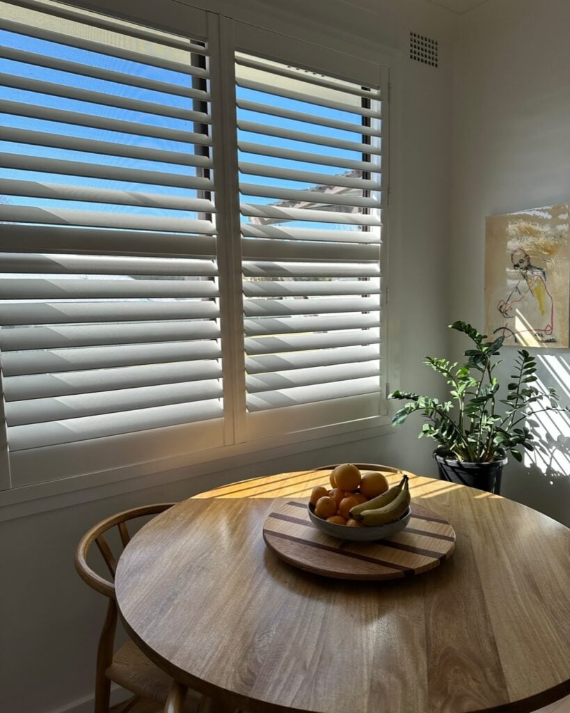 A round timber table is positioned under a window fitted with white plantation shutters, allowing strong sunlight and blue sky to stream through the slats.