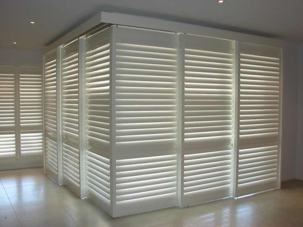 A large corner window or door opening completely covered by white bi-fold plantation shutters.