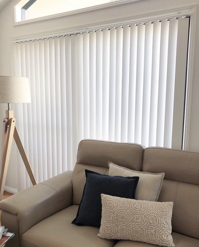 A large window is covered by white vertical blinds, and a timber tripod floor lamp stands to the side.