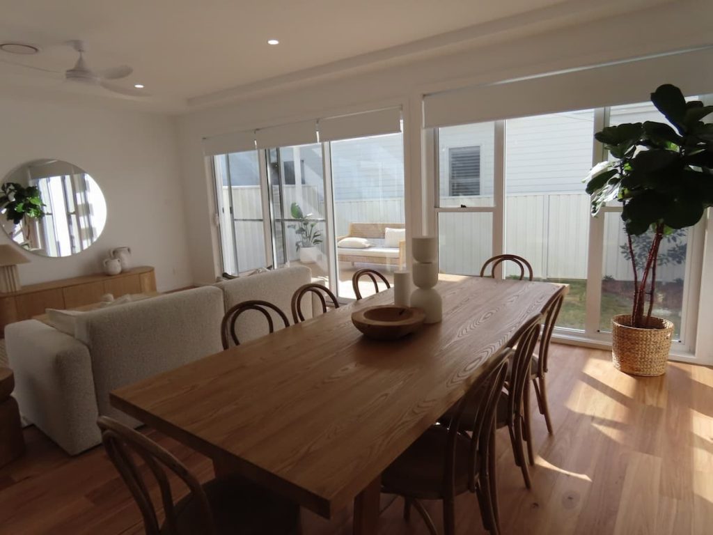 The dining area overlooks a glass sliding door and a large window, both fitted with white roller blinds and leading to an outdoor courtyard.