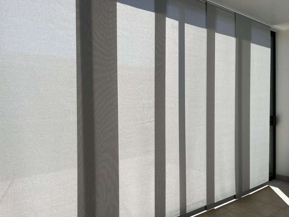 A large window or glass door covered by a track system of light grey panel glide blinds.