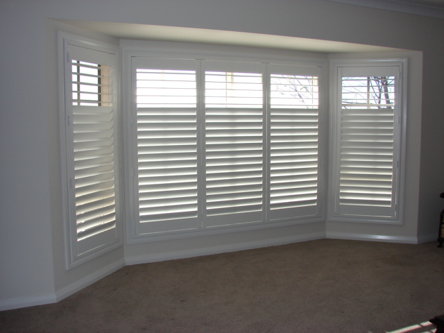 A wide bay window opening, completely fitted with white plantation shutters.