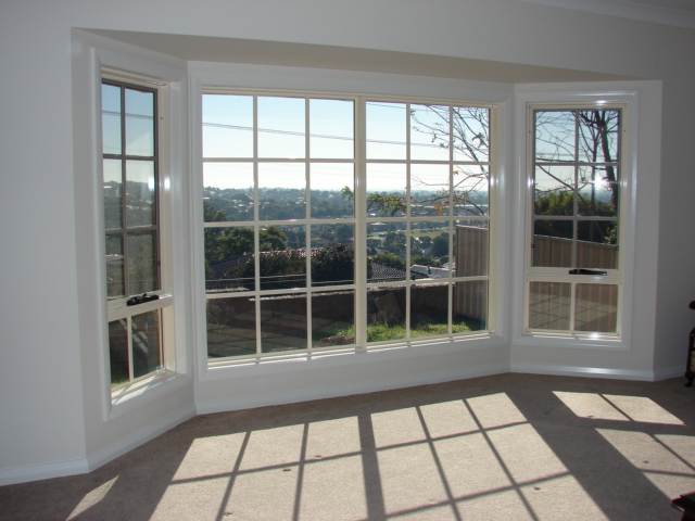 A wide, multi-panel bay window offering a sunny, elevated view of a suburban area with green trees and houses in the distance.