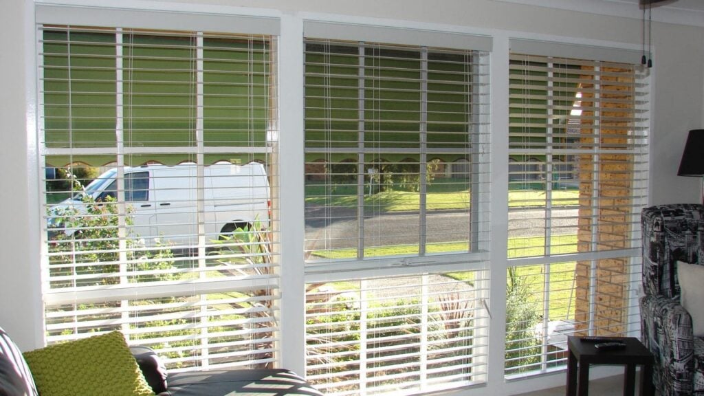 A wide, triple-panel window set with white timber frames and pale timber Venetian blinds offering a view of a suburban streetscape where a white van is parked.